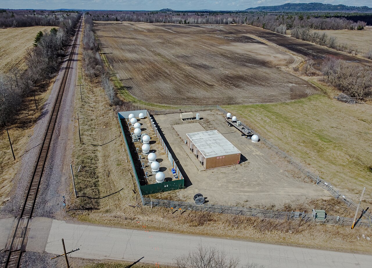 An aerial view of a fenced compound with rows of white dome-covered antennas beside farm fields and a railway.