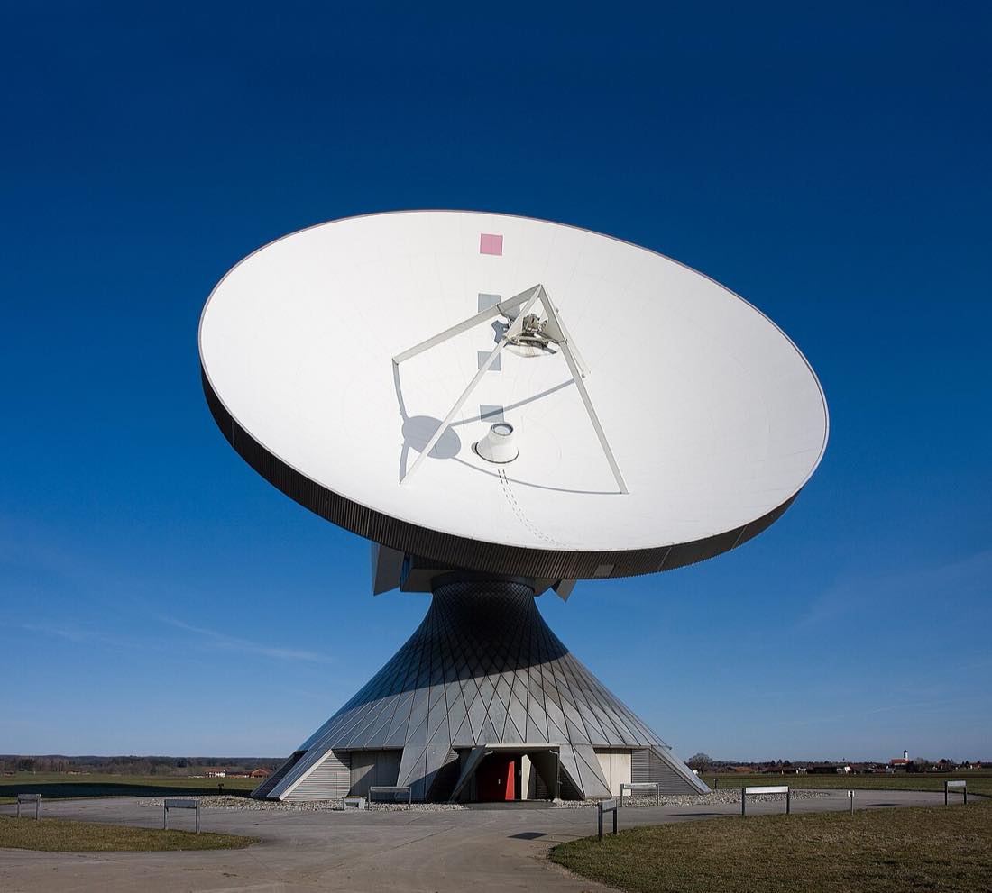 A large white satellite dish on a cone-shaped base under a blue sky.